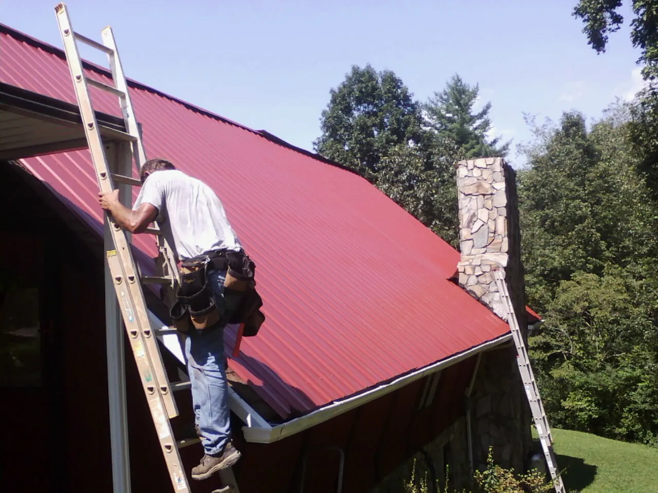 Roofer on a ladder beside a red metal roof and stone chimney — Soffit & Fascia Repair by Roofing Brothers in Cromwell